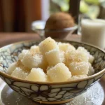 ['Cuscuz de tapioca doce com coco cremoso, em close-up.', 'Prato de cuscuz de tapioca doce com coco, mostrando textura e ingredientes.', 'Detalhe do cuscuz de tapioca doce, com pedaços de coco ralado.', 'Cuscuz de tapioca doce servido, com leite de coco por cima.']