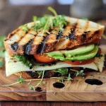 ['Panini de abacate com legumes grelhados visto de lado.', 'Close-up de um Panini de abacate e vegetais grelhados.', 'Fatias de Panini de abacate com recheio de legumes grelhados.', 'Panini com abacate, queijo e espinafre em uma apresentação apetitosa.']