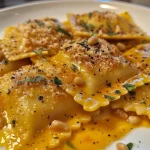 ['Ravioli de abóbora cremoso servido em um prato.', 'Close-up de ravioli recheado com abóbora e coberto com molho.', 'Prato de ravioli de abóbora com parmesão e nozes tostadas.', 'Ravioli cremoso de abóbora com ervas e queijo.']